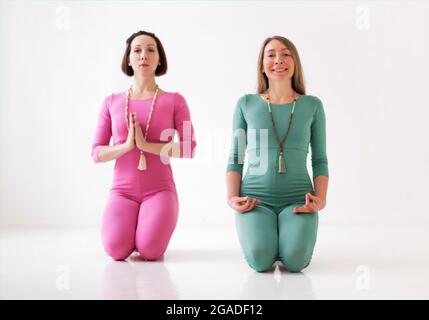 Zwei junge, glückliche, gesunde Frauen in langen Sportanzügen, die zusammen Yoga-Asanas während eines ziemlich meditativen Trainings oder einer Gruppe Yoga-Klasse durchführen, isoliert Stockfoto