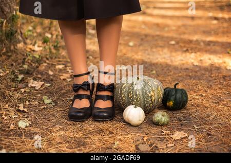 Frohe Halloween! Beschnittenes Foto von weiblichen Füßen in schwarzen Schuhen aus Retro-Leder mit Schleifen, die auf dem Ertrinkungspfad aus trockenen Blättern und Gras neben Stockfoto