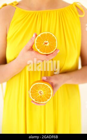 Zugeschnittenes Studiofoto eines unbekannten Weibchens in gelbem Sommerkleid mit offenen Schultern, die in den Händen zwei Hälften Orangen halten, vertikale Aufnahme. Gesund am lebendigsten Stockfoto