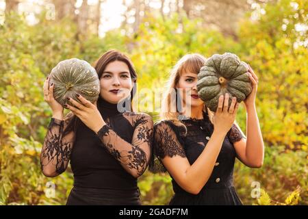 Zwei Freundinnen halten gruselige grüne Kürbisse vor ihrem Gesicht und stehen isoliert über dem herbstlichen Waldhintergrund. Herbstferien, halloween Decora Stockfoto