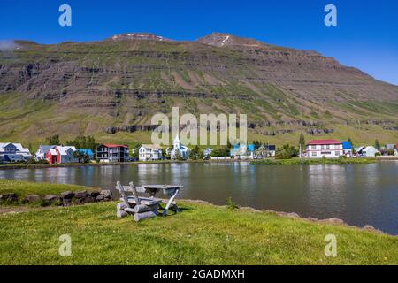 Ein rustikaler Tisch und Stuhl überblicken die hübsche Hafenstadt seydisfjordur, die sich an einem sonnigen Tag in der ruhigen Lagune widerspiegelt Stockfoto