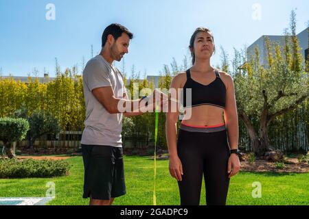 Ein europäischer Mann, ein persönlicher Trainer, misst die Hand des Klienten, die Hüfte für den Fortschritt im Training, für die Gewichtsabnahme. Im Sommer im Garten mit Pool. Stockfoto