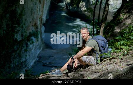 Tourist mit Rucksack, der sich ausruht, auf den Felsen sitzt und durch ein Fernglas auf eine Berglandschaft blickt. Stockfoto