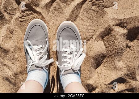 Beine in grauen Sneakers am Strand Sand. Sommerurlaub Konzept am Meer. Lifestyle-Reisen. Pov-Ansicht. Speicherplatz kopieren. Stockfoto