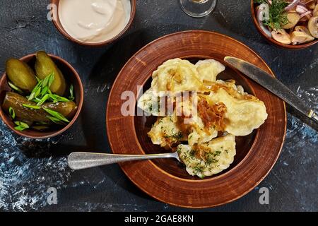 Vareniki mit Kartoffelfüllung und gebratene Zwiebeln serviert mit saurer Creme, Gurken Stockfoto