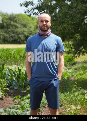 Weißer Mann in einem blauen T-Shirt und Shorts. Ein kahler Mann mit Stoppeln im Gesicht hält seine Hände in seinen Taschen. Blick in die Kamera. Stockfoto