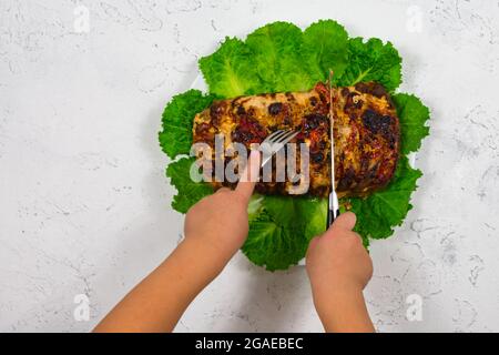 Kinderhände schneiden mit einem Messer und halten mit einer Gabel ein großes Stück gehacktes Schweinefleisch, im Ofen gekocht, im Ofen gebacken, auf Grün Stockfoto