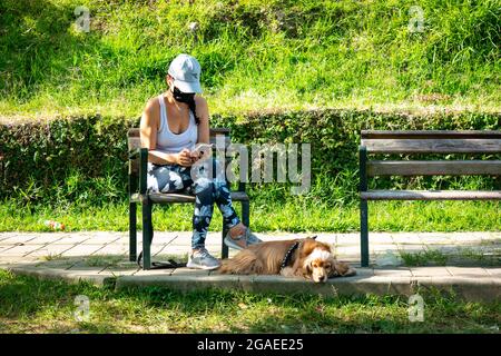 Medellin, Antioquia, Kolumbien - Juli 23 2021: Eine Lateinerin sitzt auf einer Bank und schaut ihr Telefon neben einem Mongrel Cocker Dog im öffentlichen Garten an Stockfoto