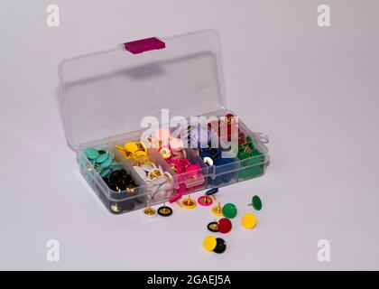 Offener Kunststoffbehälter mit zehn verschiedenfarbigen Reißnadeln in separaten Teilern mit einigen verstreuten Stiften auf weißem Hintergrund Stockfoto