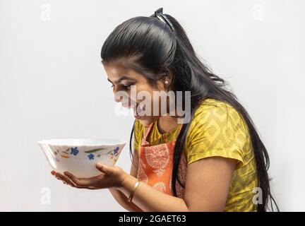 Eine hübsche indische Hausfrau in Kochschürze, die auf einen Servierkopf in der Hand auf weißem Hintergrund schaut Stockfoto