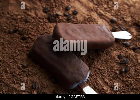 Zwei Schokoladenpopsicles auf einem strukturierten braunen Hintergrund aus Kakaopulver und Kaffeebohnen. Süßes Eis-Konzept für Design. Stockfoto