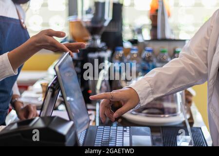 Eine junge Frau, die ein Café und einen Barista besitzt, nimmt mit einem Tablet Bestellungen von einer Kundin entgegen, die allein im Laden sitzt. Selbstentlastungsgefühl Stockfoto