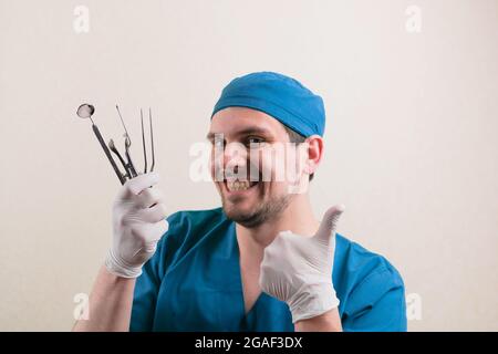 Junger lächelnder Arzt mit medizinischen Instrumenten in der Hand und Daumen hoch in blauem Chirurgenanzug isoliert Stockfoto