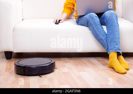 Mann, der an der Fernarbeit mit Laptop arbeitet, während der Roboter-Staubsauger das Wohnzimmer und den Staub reinigt. Zeit für sich selbst. Mann steuert den vac Stockfoto