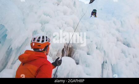Mann steht und kontrolliert ein Sicherheits-Top-Seil, während Weibchen mit Eiskletterausrüstung auf einem gefrorenen Wasserfall klettert Stockfoto
