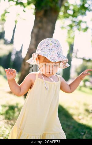 Das kleine Mädchen in einem panamahut geht entlang des Rasens, hebt ihre Arme und schaut auf ihre Füße Stockfoto