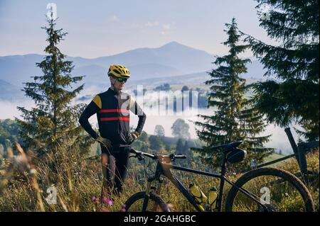Radfahrer in Uniform und Helm, stehend auf einem grasbewachsenen Hügel. Schöne Berglandschaft im Hintergrund. Am frühen Morgen in den neblig sonnigen Bergen. Fahrrad auf verschwommenem Vordergrund. Stockfoto