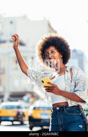Junge Afro-Frau, die auf der Straße steht und den Arm hebt, um auf ein Taxi aufmerksam zu machen. Sie hält ein Smartphone auf der anderen Hand. Konzept der Taxi-Anwendung Stockfoto