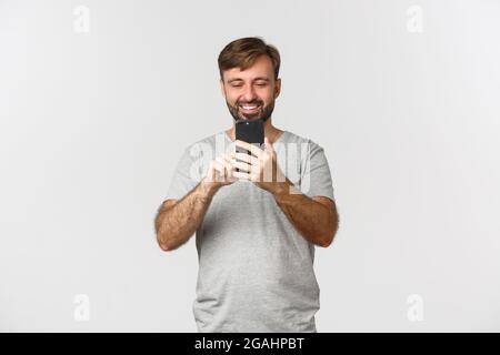 Porträt eines erwachsenen Mannes mit Bart, trägt graues T-Shirt, Foto auf dem Smartphone oder Aufnahme eines Videos, stehend auf weißem Hintergrund Stockfoto