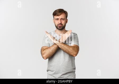 Enttäuschter Mann mit Bart, zeigt Kreuzgesten und Stirnrunzeln, sagt zu stoppen, verbieten etwas Schlechtes, stehen über weißem Hintergrund Stockfoto