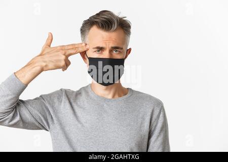 Konzept von covid-19, sozialer Distanzierung und Quarantäne. Nahaufnahme eines traurigen Mannes mittleren Alters in schwarzer medizinischer Maske, mit der Fingerpistole auf den Kopf zeigend und schauend Stockfoto