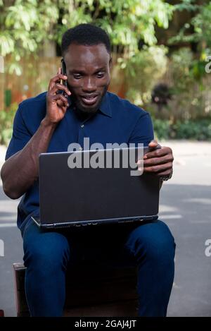 Junger Mann, der im Freien sitzt, mit dem Mobiltelefon kommuniziert und lächelnd auf den Laptop schaut. Stockfoto