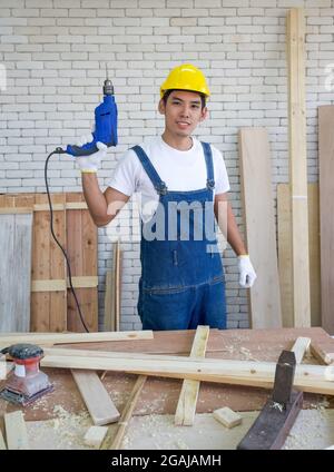 Asiatische Tischler trägt einen gelben Hardhut, stellt sich selbstbewusst vor Beginn der Arbeit der Annahme von Aufträgen am Holzarbeitsplatz. Morgenarbeit atmos Stockfoto