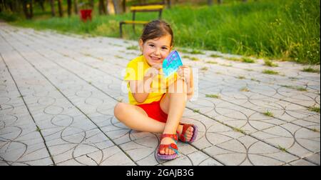 Fröhliches kleines Mädchen auf dem Weg im Park sitzen Kreuz Beine halten in ihren Händen und Stress Spielzeug bunt Pop it und mit lächelndem Gesicht Blick auf die Stockfoto