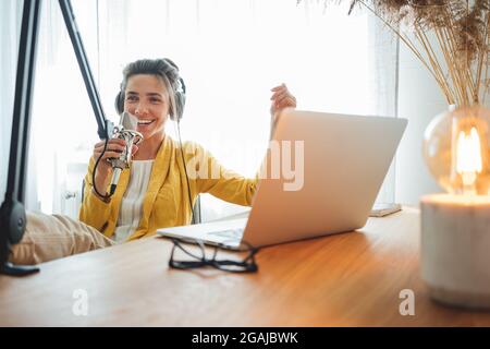 Fröhliche Frau Podcasterin, die ihre Stimme ins Mikrofon aufgenommen hat. Radiohosten streamen Podcast mit Mikrofon und Laptop in seinem Heimstudio Stockfoto