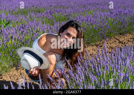 Eine junge Frau kauerte sich in einem blühenden Lavendelfeld. Sie hat gerade ihren Hut abgenommen und lächelt glücklich. Lässiger Stil und lange braune Haare. Stockfoto