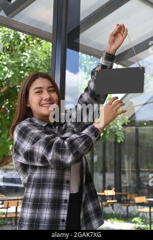 Lächelnde Frau, die ein leeres Holzschild an der Glastür hing. Für Ihren Werbetext. Stockfoto