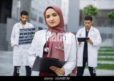 Nahaufnahme einer weiblichen muslimischen angenehmen Ärztin in Hijab, die mit Klemmbrett vor dem Krankenhausgebäude steht, während zwei hübsche arabische Ärzte hinter sich mit Röntgen- und Tablet-pc arbeiten Stockfoto