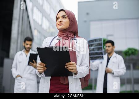 Ziemlich lächelnde arabische Ärztin in Hijab posiert draußen vor der modernen Klinik und schaut weg. Zwei selbstbewusste männliche Ärzte, die hinter sich stehen und mit MRI-Scans und Tablet-pc arbeiten Stockfoto