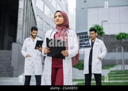 Ziemlich lächelnde arabische Ärztin in Hijab posiert draußen vor der modernen Klinik und schaut weg. Zwei selbstbewusste männliche Ärzte, die hinter sich stehen und mit MRI-Scans und Tablet-pc arbeiten Stockfoto