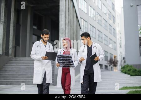 Drei Ärzte, eine Frau im Hijab und zwei Männer in medizinischer Kleidung, diskutierten über die Röntgentomographie des Patienten und gingen mit einer Treppe nach draußen auf den Hintergrund eines modernen Krankenhauses Stockfoto