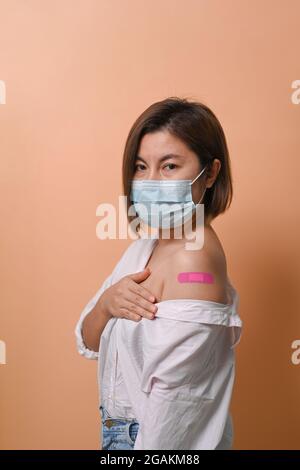 Asiatische Frau, die auf beigem Hintergrund steht und ihren Arm zeigt, nachdem sie einen Impfstoff erhalten hat. Stockfoto