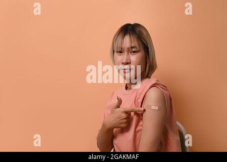 Asiatische Frau sitzt auf beigem Hintergrund und zeigt ihren Arm mit Verband, nachdem sie eine Impfung erhalten hat. Stockfoto