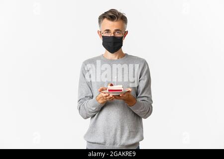 Konzept von Coronavirus, Quarantäne und Urlaub. Bild von traurigen Mann mittleren Alters in schwarzer medizinischer Maske und Brille, hält Geburtstagskuchen mit brennenden Kerze Stockfoto