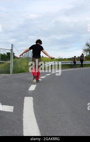 Lockiger Teenager, der auf den Schultermarkierungen auf der Straße balanciert. Von hinten gesehen, während man in einem lagermarsch läuft, vor ihm ist ein Mädchen und ein Erwachsener wo Stockfoto