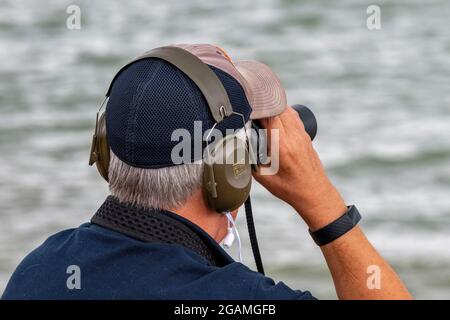 Älterer Mann mit Fernglas, Rentner, der durch ein Fernglas schaute, Mann mit Fernglas, Mann mit Ohrenschützern, Zuschauer bei der cowes Week, Spionage. Stockfoto