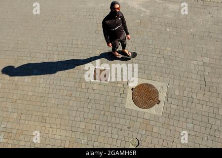 Junger erwachsener Mann mit Sonnenbrille und Maske, der auf einem Skateboard läuft, von oben gesehen. Handspuren im Zement um den Metallablauf. Stockfoto