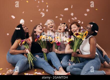 Eine Gruppe von verschiedenen Frauen mit Blumensträußen, die unter fallenden Blütenblättern Spaß haben. Sechs glückliche Weibchen sitzen zusammen auf einem braunen Hintergrund. Stockfoto