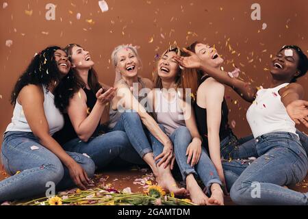 Gruppe von sechs lachenden Frauen unterschiedlichen Alters, die unter fallenden Blütenblättern sitzen. Multi-ethnische lächelnde Frauen, die Spaß im Studio haben, während sie auf sitzen Stockfoto