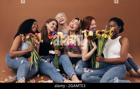 Sechs Frauen unterschiedlichen Alters und Körpertypen halten Blumensträuße, während sie im Studio vor einem braunen Hintergrund sitzen Stockfoto