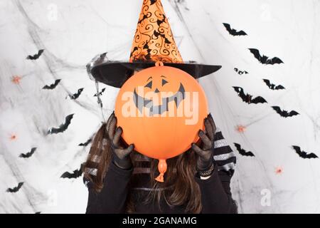 Eine Frau in einem Hexenkostüm in einem Hut hält einen Kugelkürbis. Halloween-Leute. Stockfoto