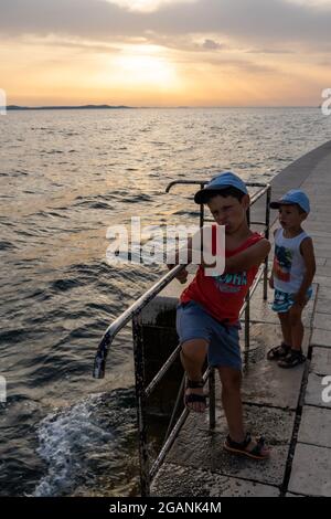 ZADAR, KROATIEN - 21. Jul 2021: Eine vertikale Aufnahme von Kindern, die bei Sonnenuntergang in Kroatien an einer Metallbarriere in der Nähe des Wassers stehen Stockfoto