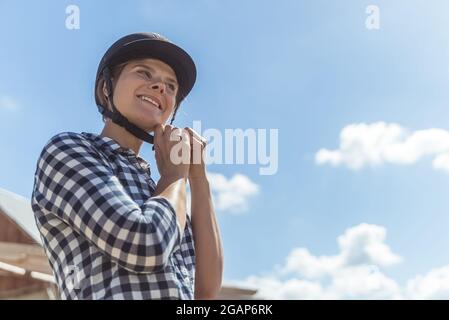 Weibliche Jockey, die einen Helm aufsetzt und sich für den Reitwettbewerb vorbereitet. Eine wettbewerbsfähige Frau ist auf den Wettbewerb eingestellt. Konzept des Reitwettbewerbs. Wolkiger Himmel im Hintergrund. Stockfoto