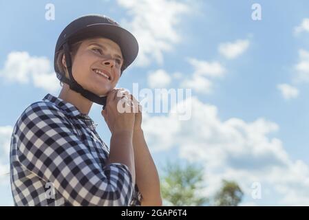 Weibliche Jockey, die einen Helm aufsetzt und sich für den Reitwettbewerb vorbereitet. Eine wettbewerbsfähige Frau, die alle auf den Wettbewerb eingestellt ist. Konzept des Reitwettbewerbs. Wolkiger Himmel im Hintergrund. Stockfoto