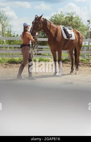 Weibliche Jockey mit ihrem dunkelbraunen Pferd in der sandigen Arena mit einem Holzzaun. Vorbereitung auf den Reitwettbewerb. Das Mädchen hält das Bleiseil des Pferdes und fixiert seinen Zaumzeug. Stockfoto