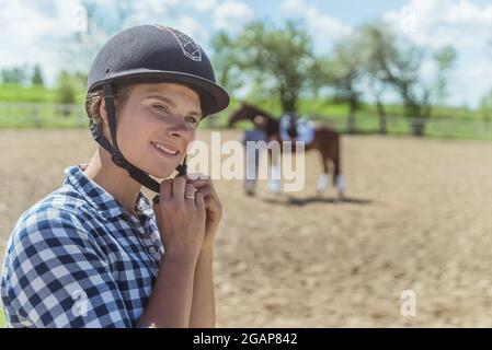 Weibliche Jockey, die einen Helm aufsetzt und sich für das Reiten vorbereitet. Ein Mädchen mit einem dunklen Lorbeer im Hintergrund. Sandiger Parkour mit einem Holzzaun. Reiten zur freien Verfügung. Stockfoto